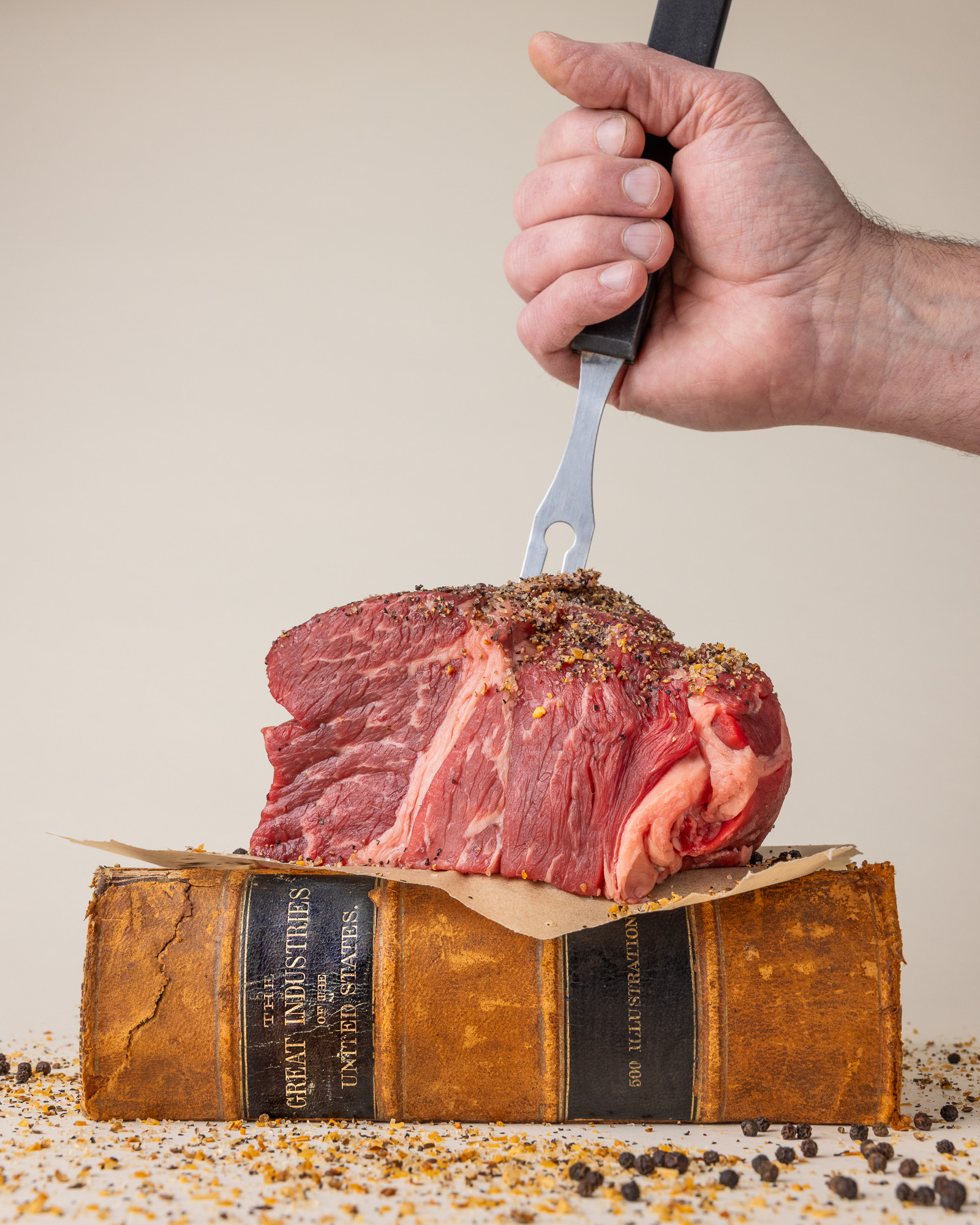 rancher's hand holding bbq fork stabbing into a raw beef roast sitting on top of parchment paper and an antique book about great industries of united states